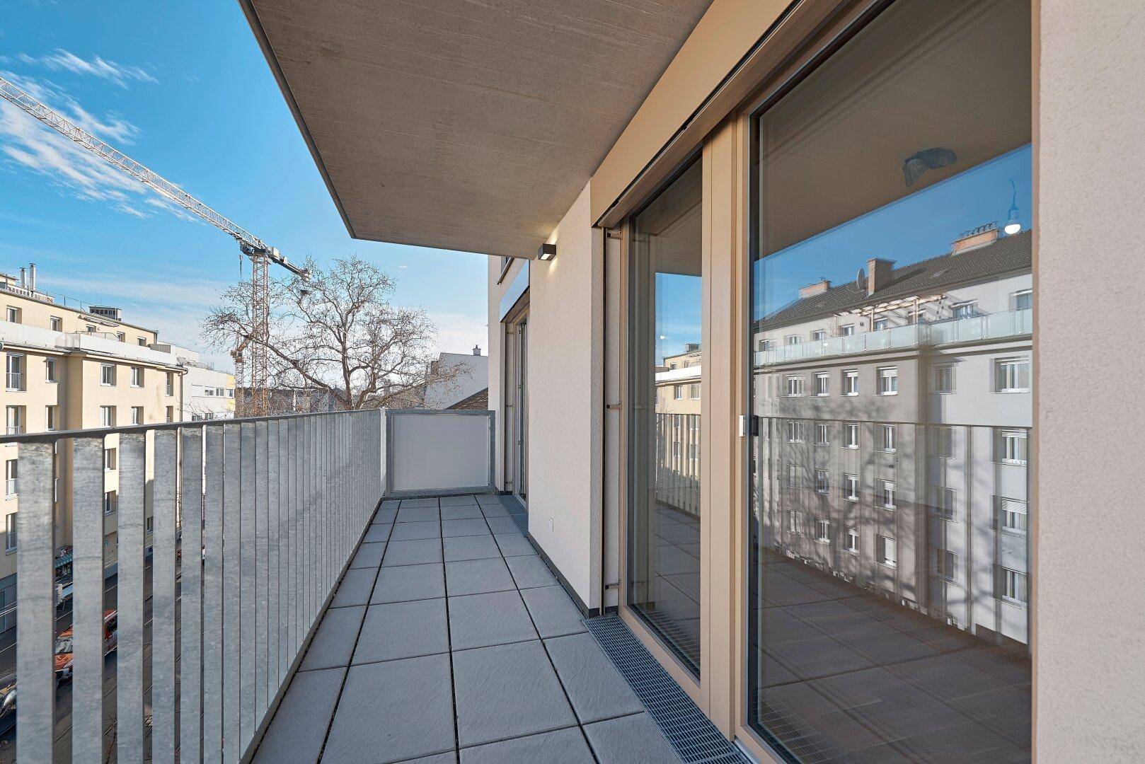 Langer Balkon mit Fliesenboden und Metallgeländer, bietet Blick auf die Stadt und umliegende Gebäude.