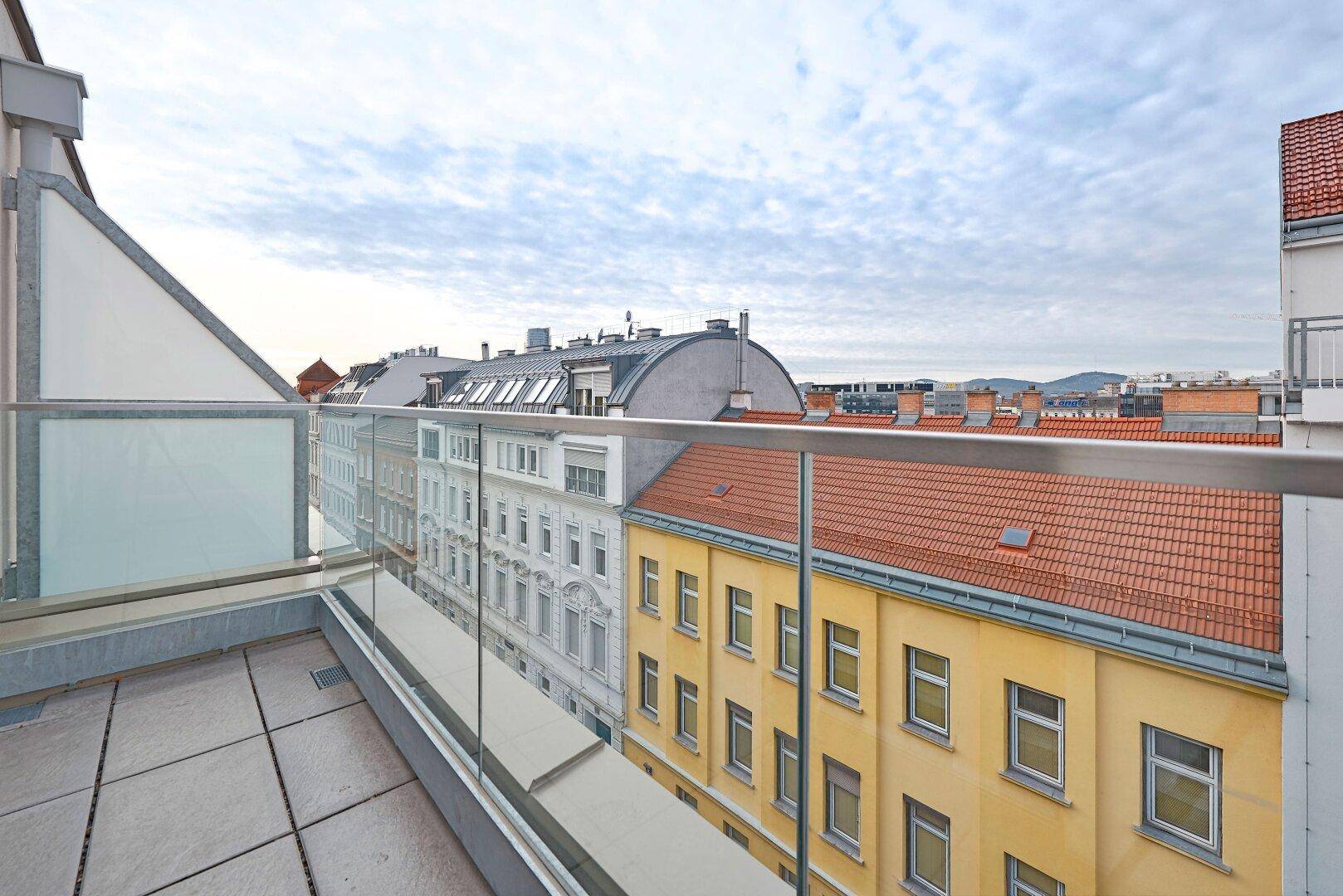 Balkon mit Fliesenboden und Glasgeländer, bietet einen Blick auf die umliegenden Gebäude.