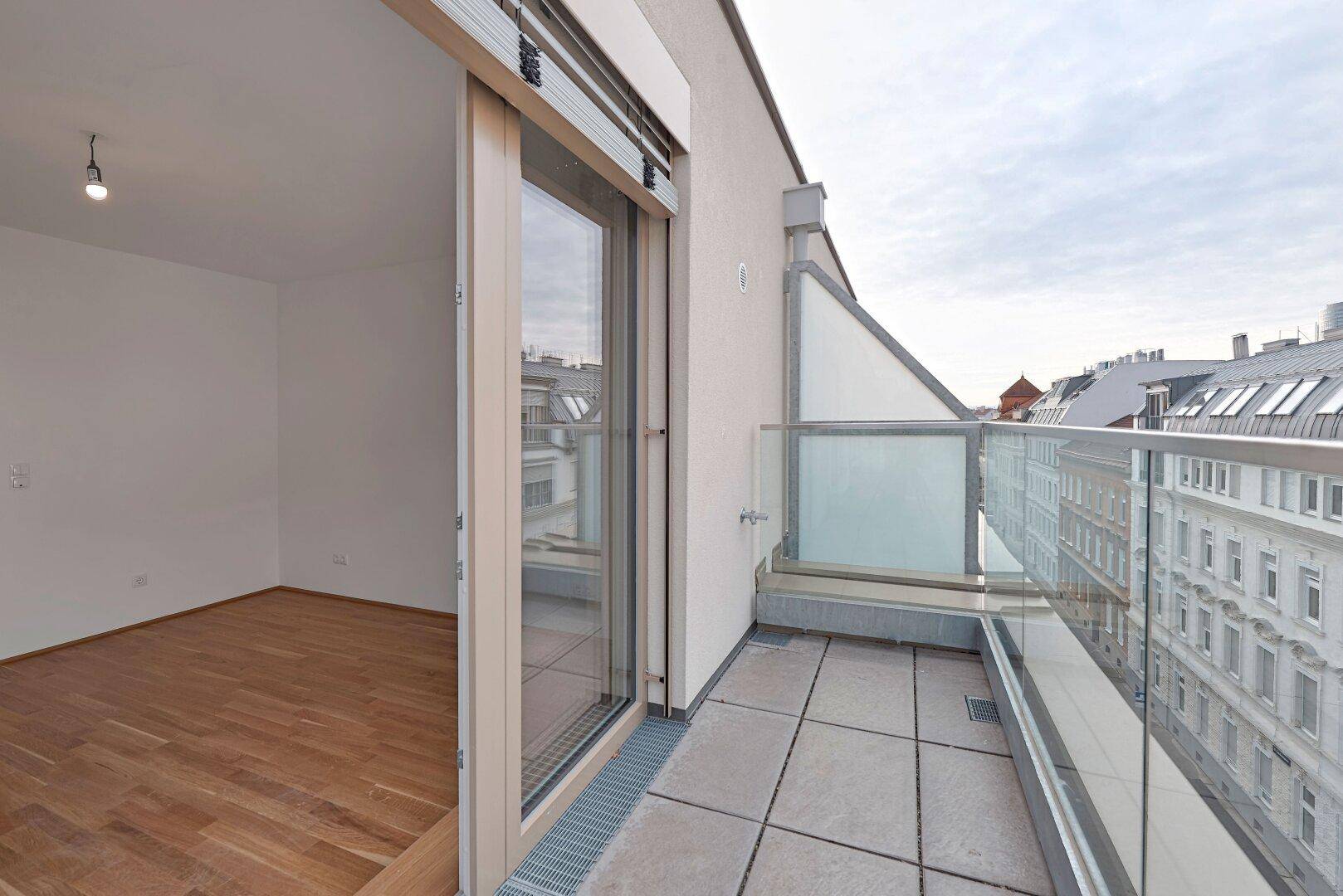 Heller Balkon mit Fliesenboden, Glasgeländer und großem Schiebetürzugang zum Innenraum.