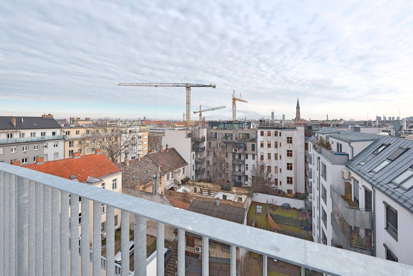 Weitblick von einem Balkon über die Stadt mit Gebäuden, Kränen und Hügeln im Hintergrund.