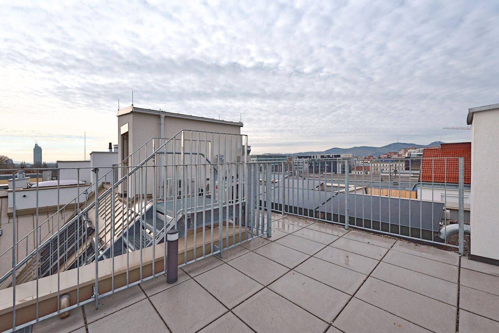 Weitläufige Dachterrasse mit Fliesenboden und Metallgeländer, bietet einen Panoramablick über die Stadt.