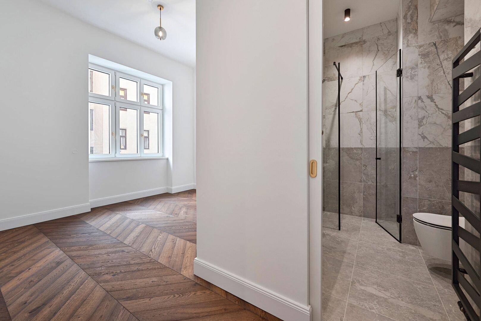 Blick von einem leeren Zimmer mit Parkettboden in ein modernes Badezimmer mit Dusche.