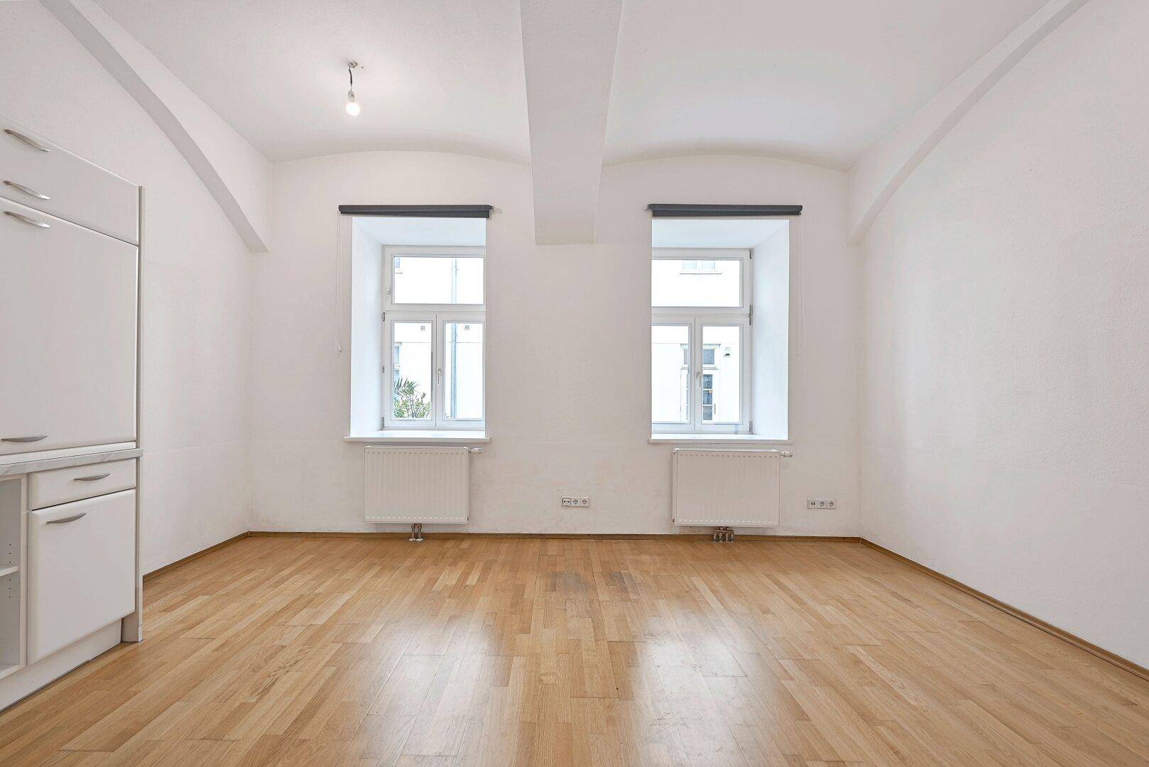 Heller Wohnraum mit zwei Fenstern, Holzboden und hohen Decken.