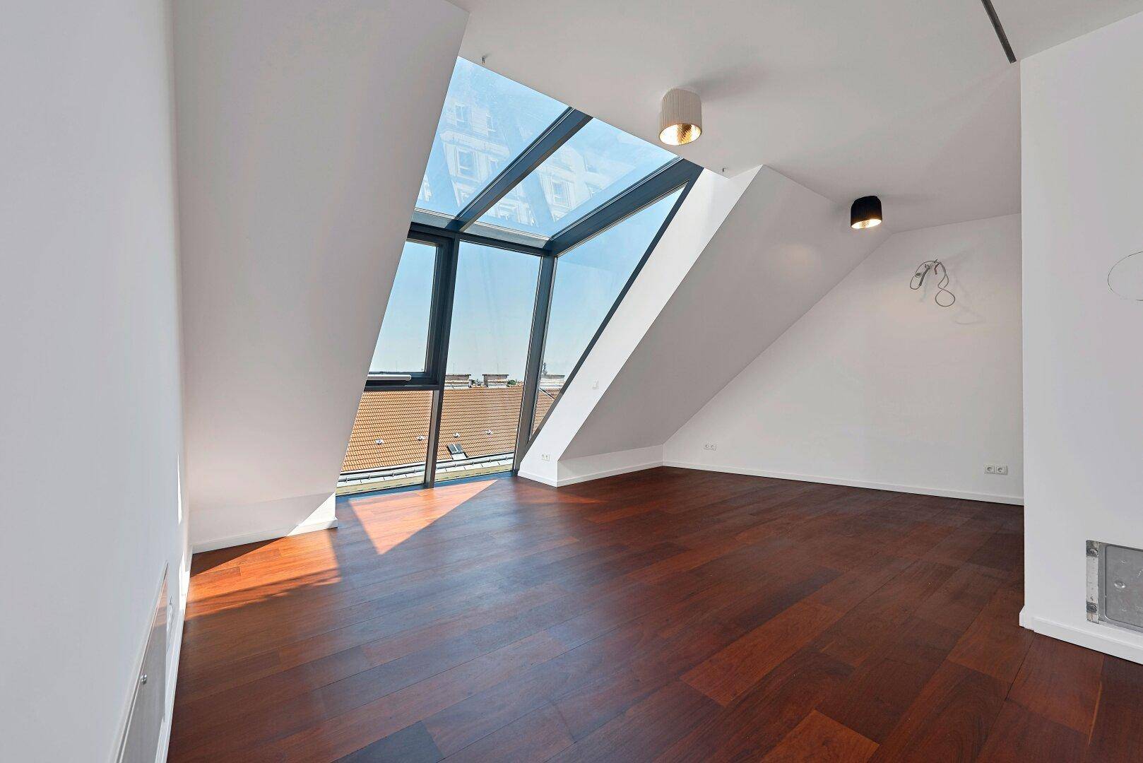 Lichtdurchfluteter Raum mit dunklem Holzfußboden, schrägen Wänden und beeindruckenden Dachfenstern.
