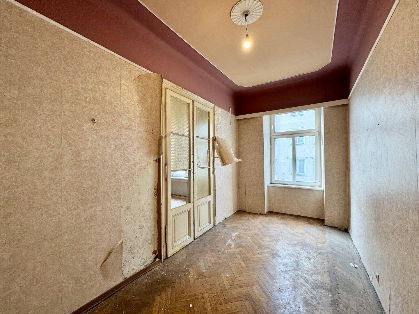 Geräumiges Zimmer mit beschädigtem Fischgrätparkett, alter Tapete und hohen Decken, bereit zur Renovierung.