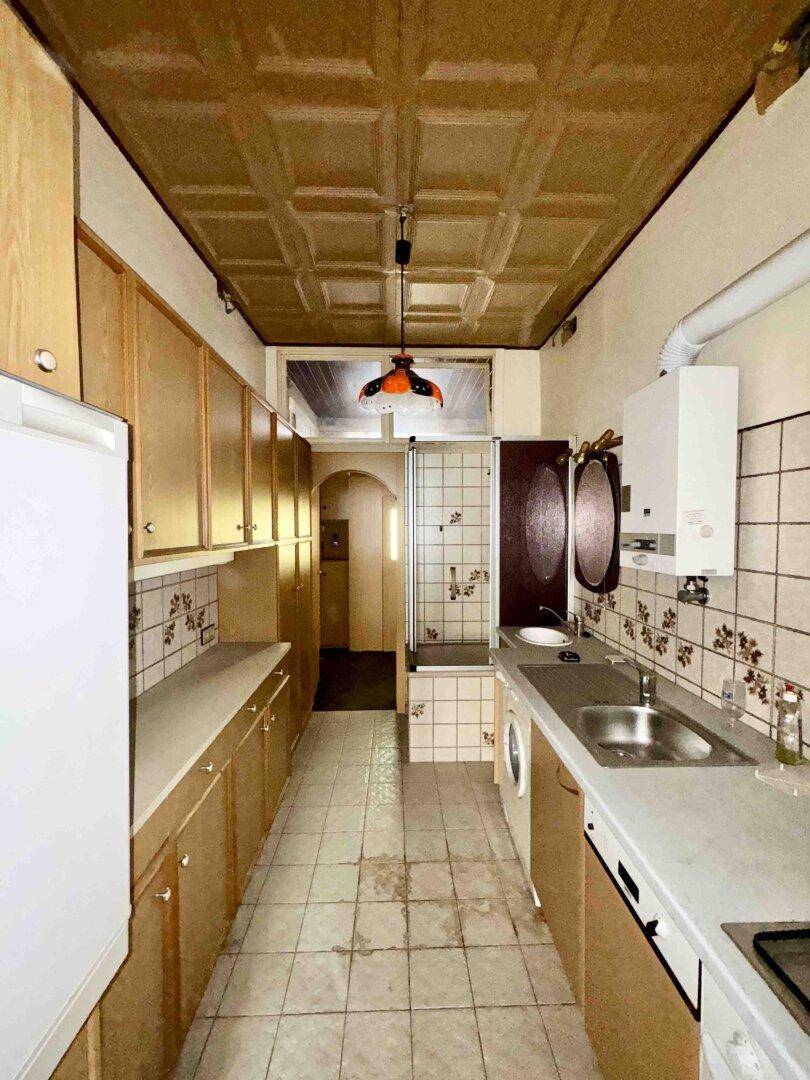 Schmale Küche mit älteren Holzschränken, Fliesenboden und Deckenpaneelen, in originalem Zustand.