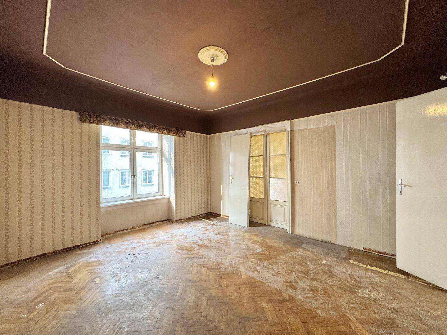 Leeres Zimmer mit abgenutztem Fischgrätparkett, alter Tapete und Deckenstuck in Renovierungszustand.