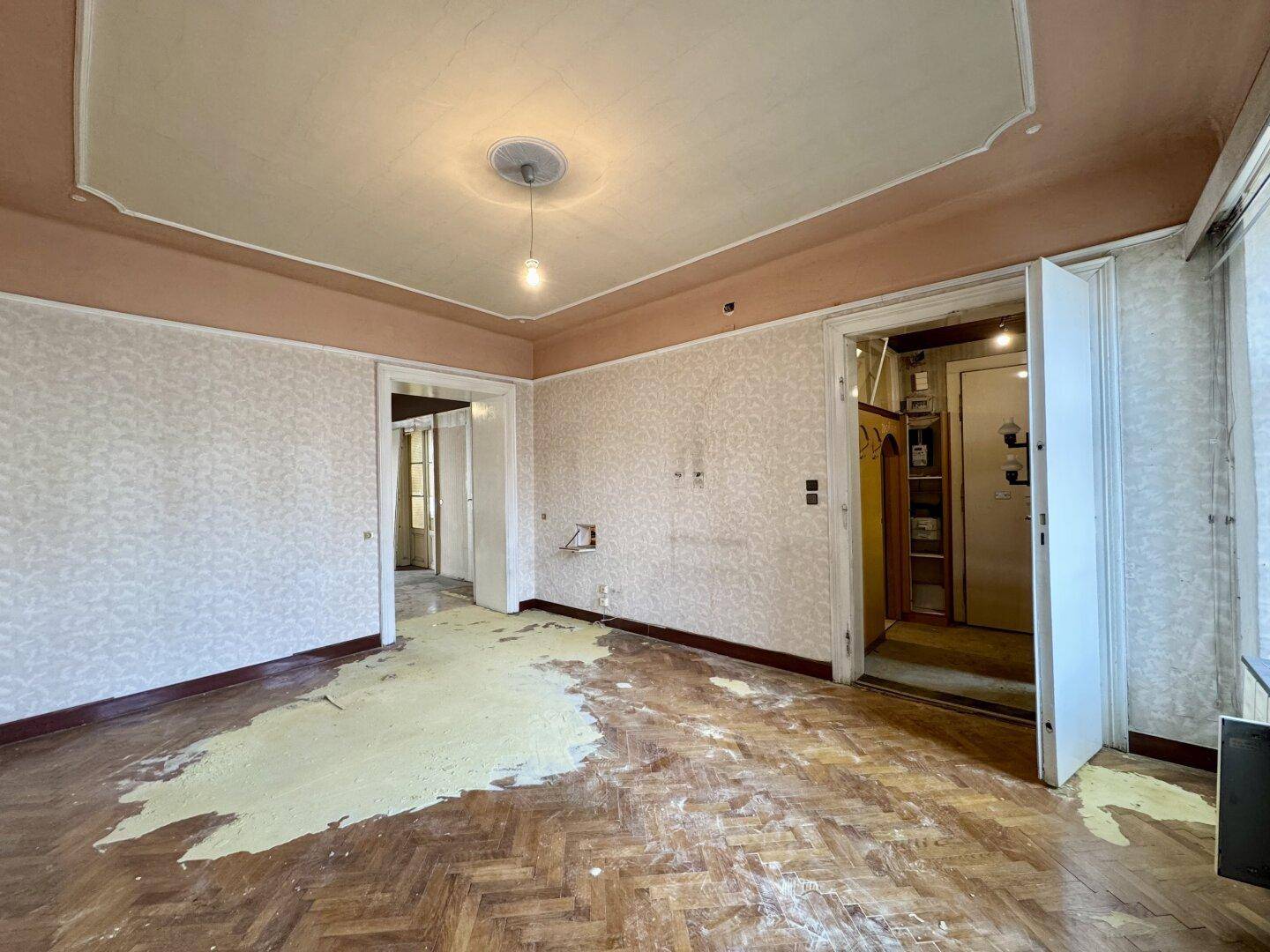 Geräumiges Zimmer mit abgenutztem Fischgrätparkett und zwei Türöffnungen, bereit zur Modernisierung.