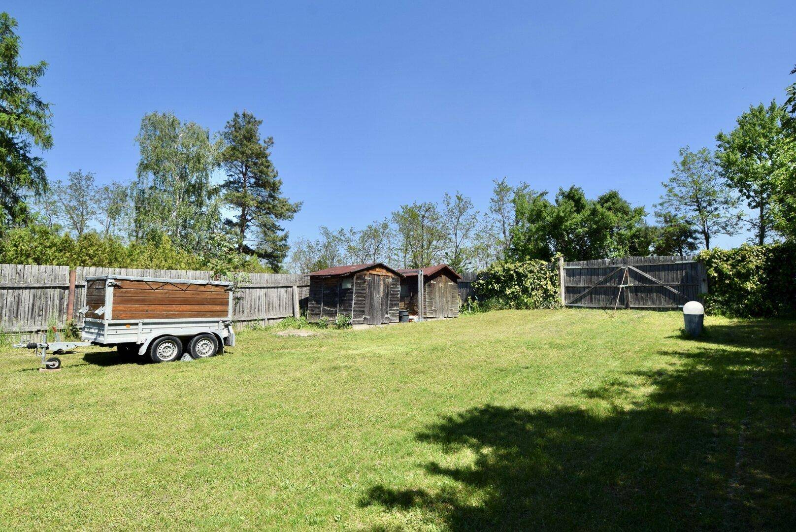 Weitläufiger Garten mit Rasenfläche, mehreren Holzschuppen und einem Anhänger, umgeben von Bäumen.