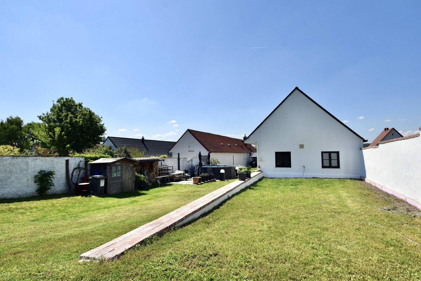 Großer Garten mit Rasenfläche und einem langen Weg, der zum Haus führt, unter blauem Himmel.