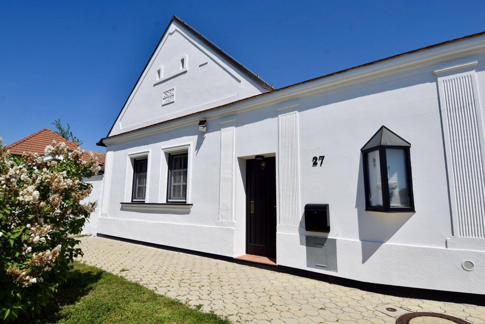 Charmante weiße Hausfassade mit schwarzen Fenstern und Haustür, umgeben von Grün.