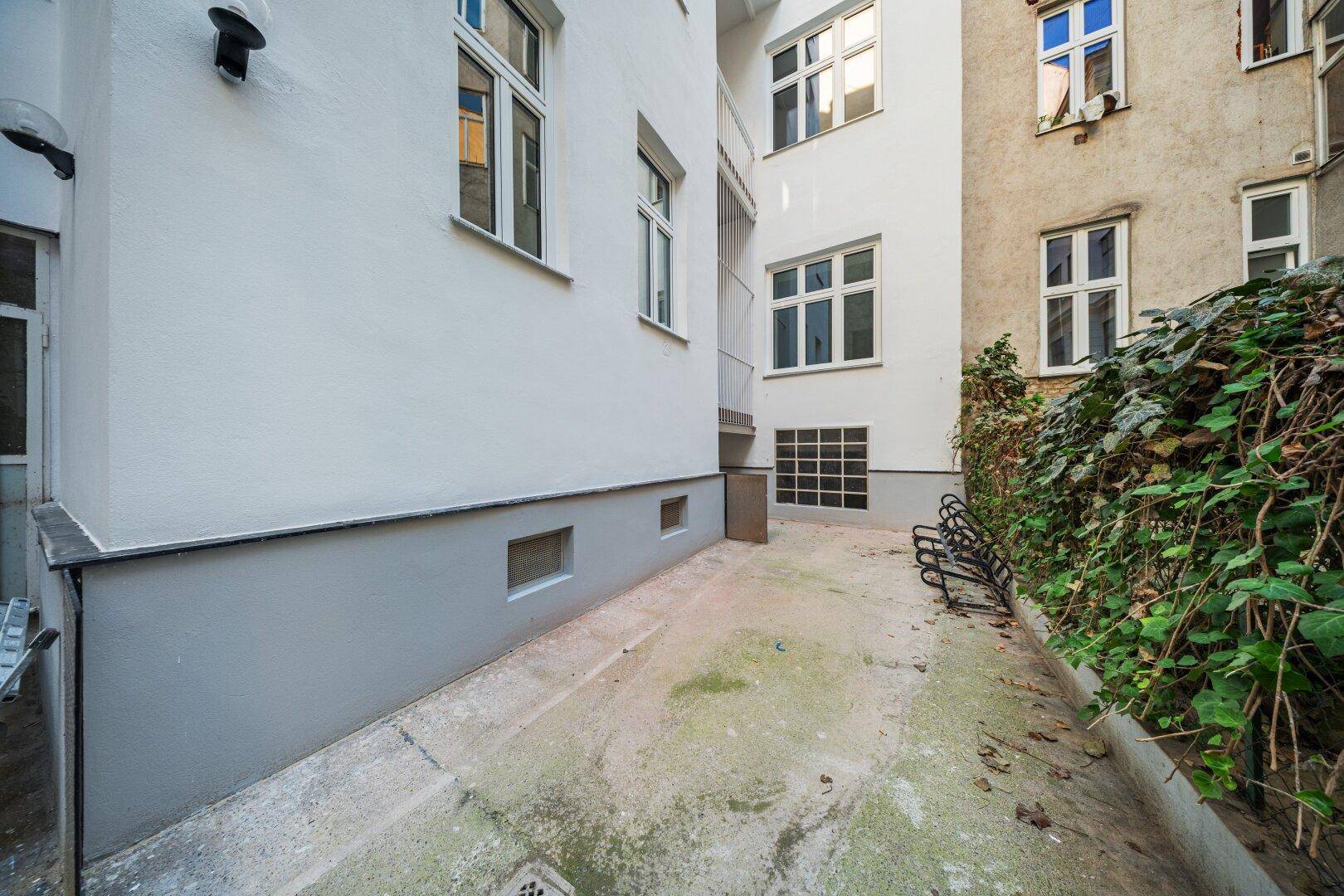 Schmaler Außenbereich zwischen Gebäuden mit Betonweg und etwas Grünbewuchs.
