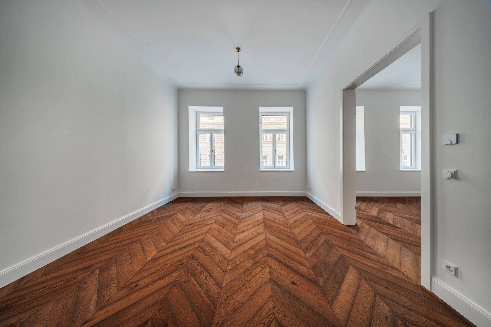 Geräumiges Zimmer mit elegantem Fischgrätparkett und zwei großen Fenstern.