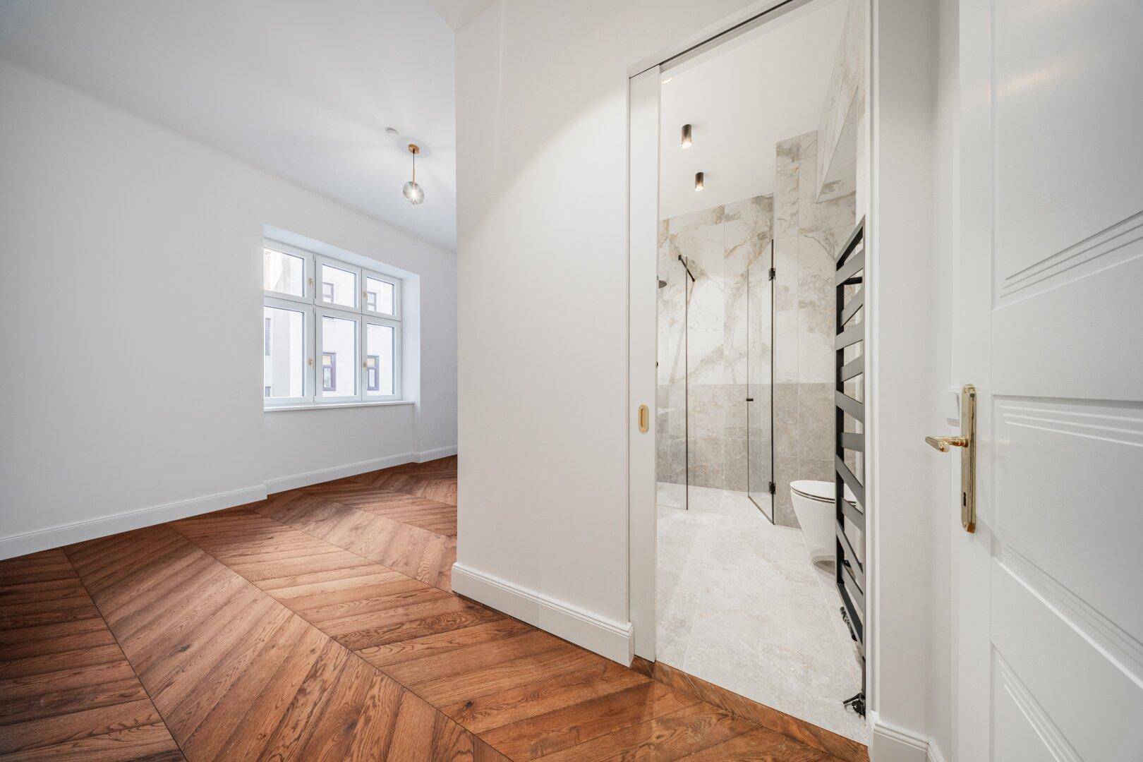 Blick vom Zimmer in einen Flur mit Fischgrätparkett und Zugang zu einem modernen Badezimmer.