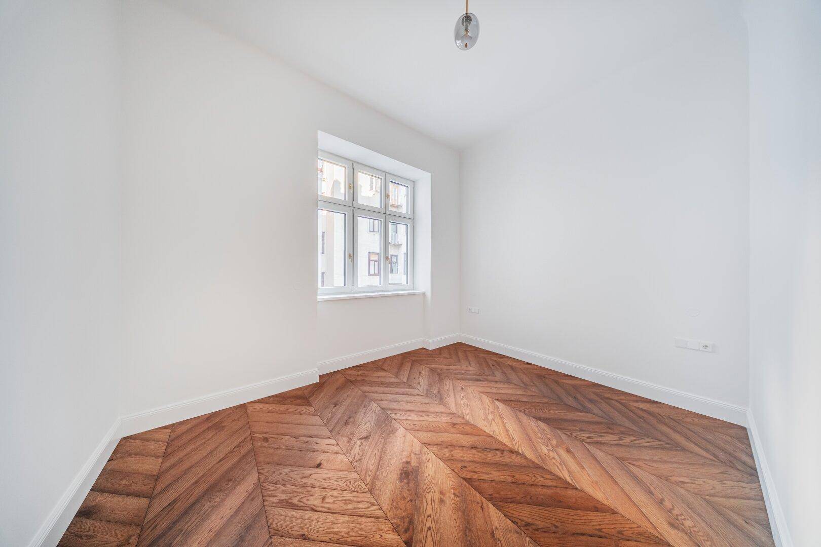 Gemütliches, helles Zimmer mit Fischgrätparkett und einem großen Fenster.