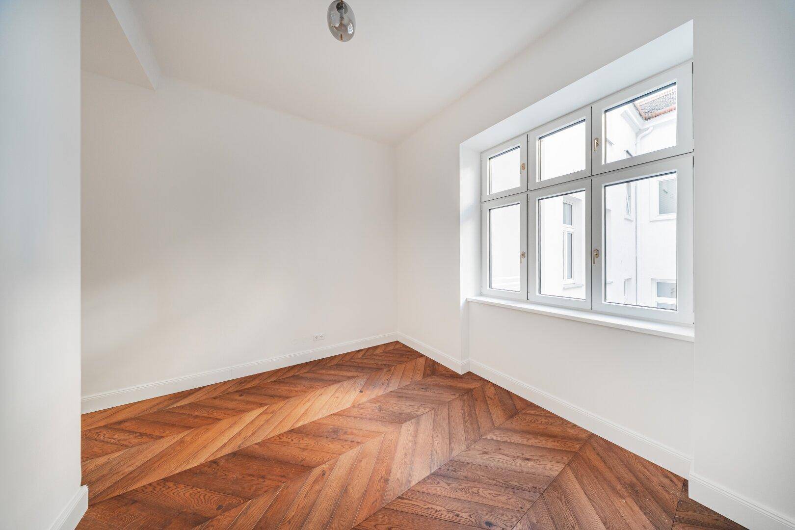 Helles Schlafzimmer mit Fischgrätparkett und einem breiten Fenster, ideal für Ruhe.