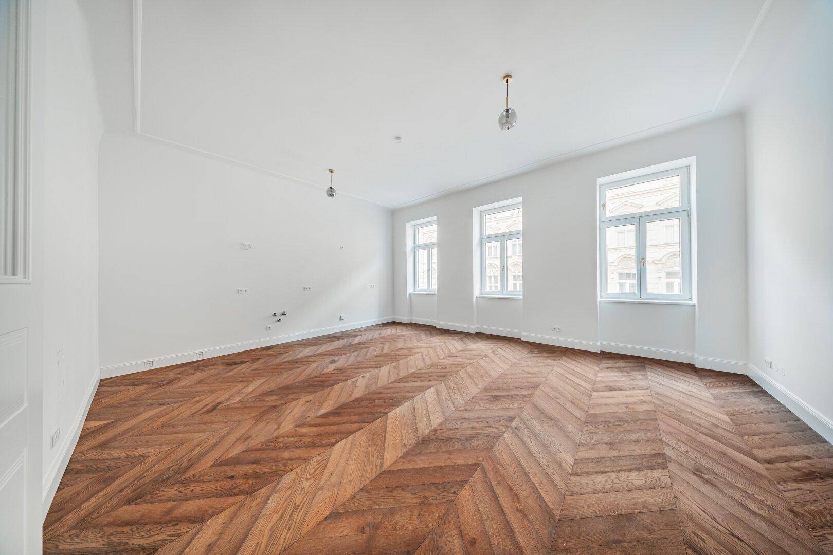 Geräumiges, helles Zimmer mit elegantem Fischgrätparkett und mehreren Fenstern.