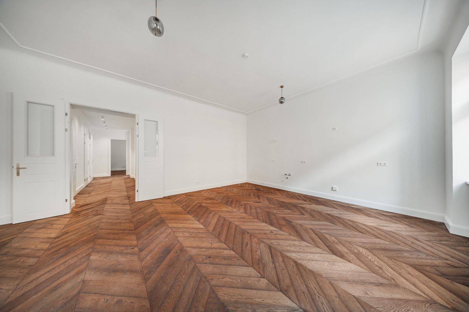 Weitläufiges Zimmer mit Fischgrätparkett und Zugang zu einem hellen Flur.