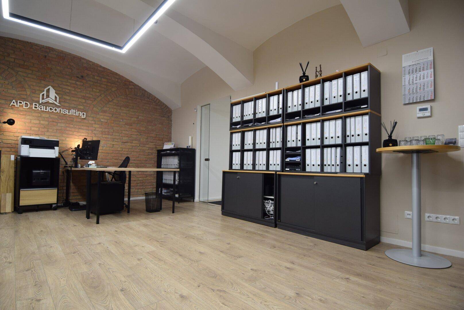 Geräumiges Büro mit modernem Design, Holzboden, Ziegelwand und gewölbter Decke, ausgestattet mit Büromöbeln und Regalen.