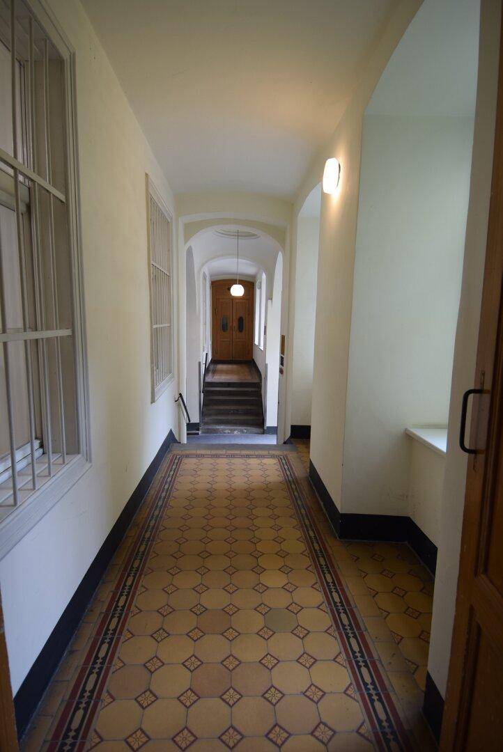 Langer Flur mit gemustertem Fliesenboden, gewölbter Decke und Treppenaufgang, mit Fenstern und Gitterstäben.