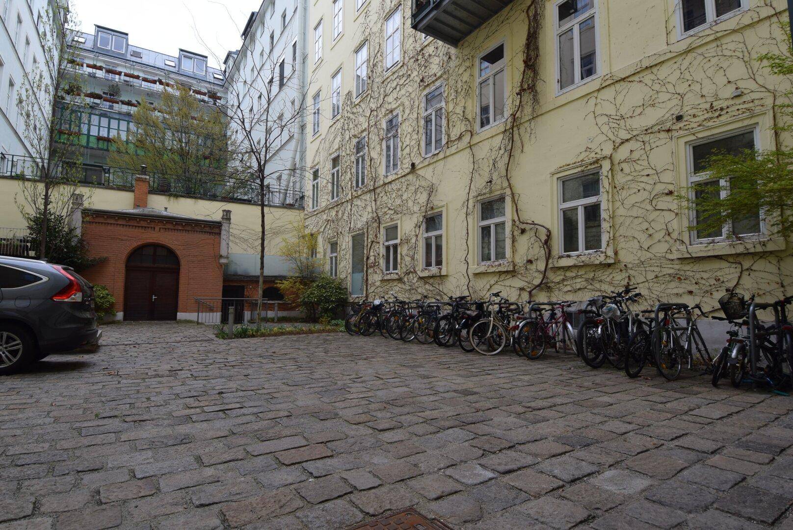 Innenhof mit Kopfsteinpflaster, einer von Efeu bewachsenen Fassade, geparkten Fahrrädern und einem Auto.