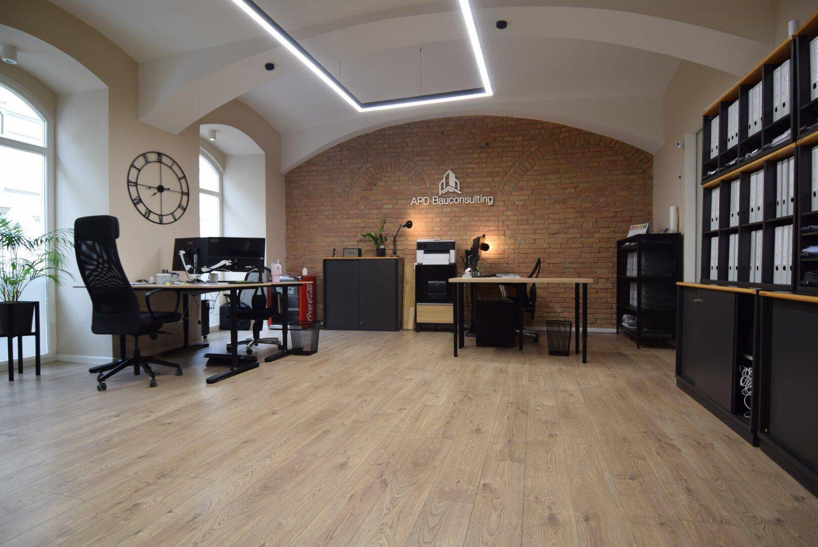 Großes Büro mit mehreren Arbeitsplätzen, Ziegelwand, gewölbter Decke und großen Fenstern, ausgestattet mit Büromöbeln.