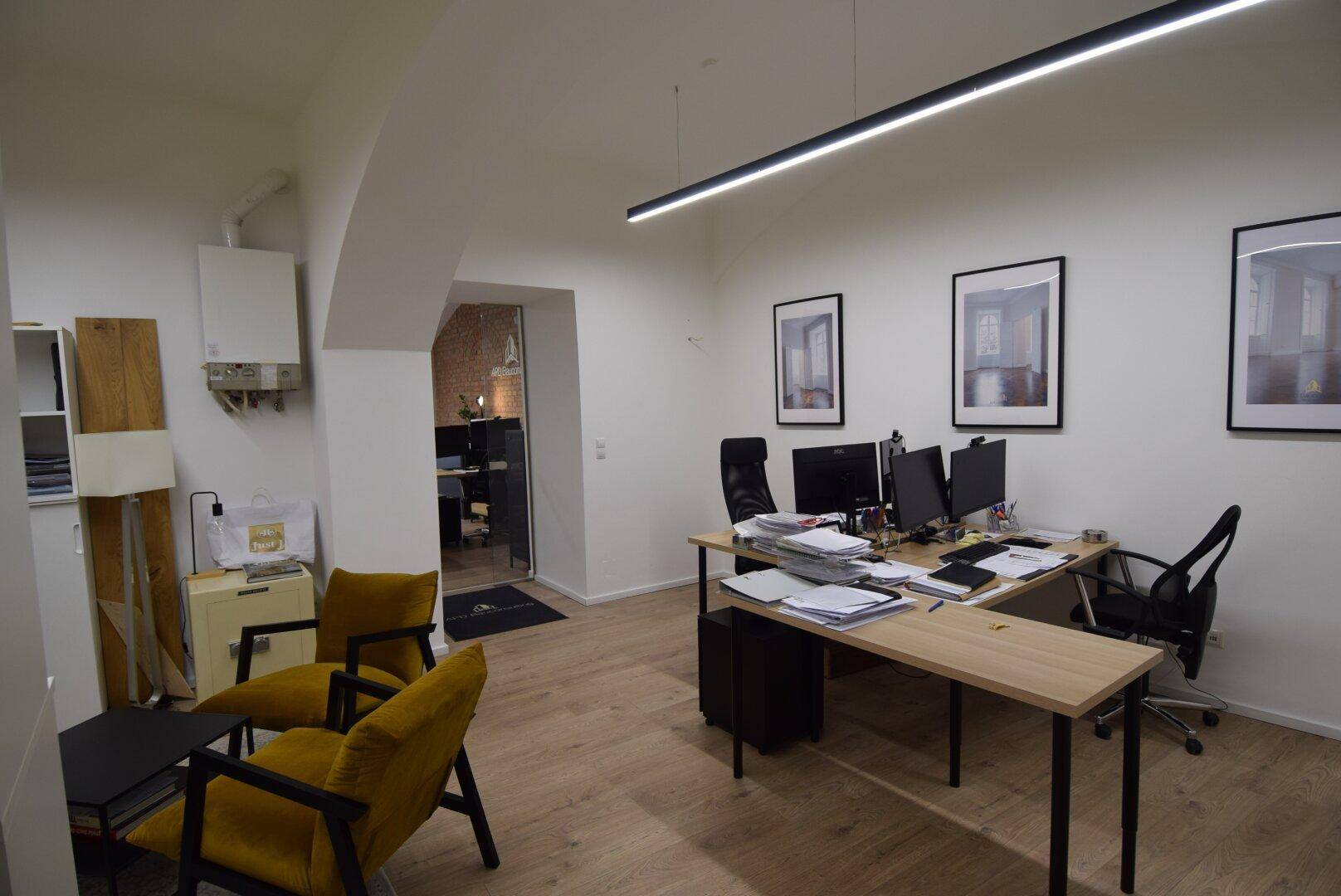Büro mit mehreren Arbeitsplätzen, gelben Sesseln und einem Wandheizkessel, mit Holzboden und weißen Wänden.