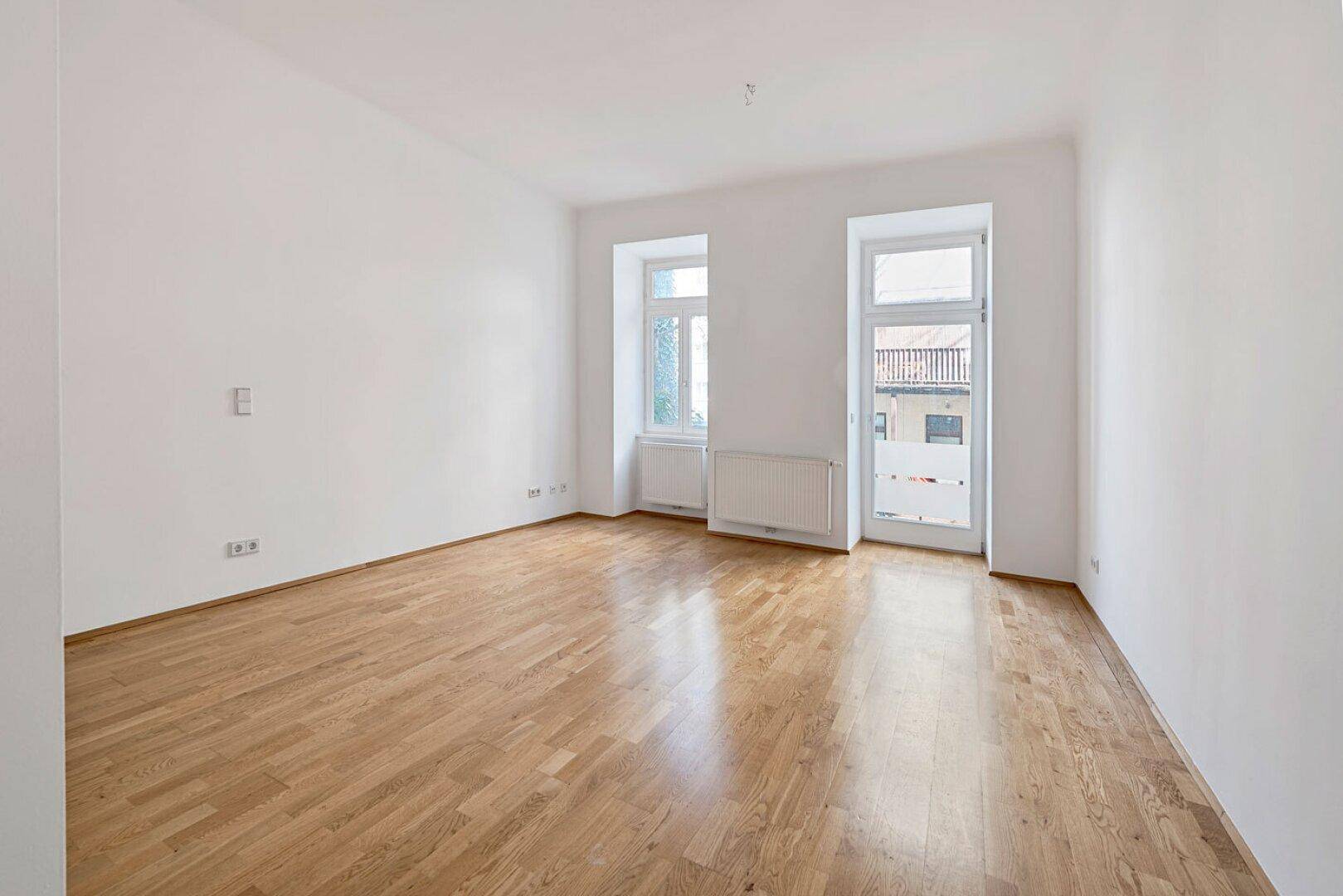 Geräumiges Zimmer mit Holzboden, weißen Wänden und Fenstern, eines davon mit Zugang nach draußen.