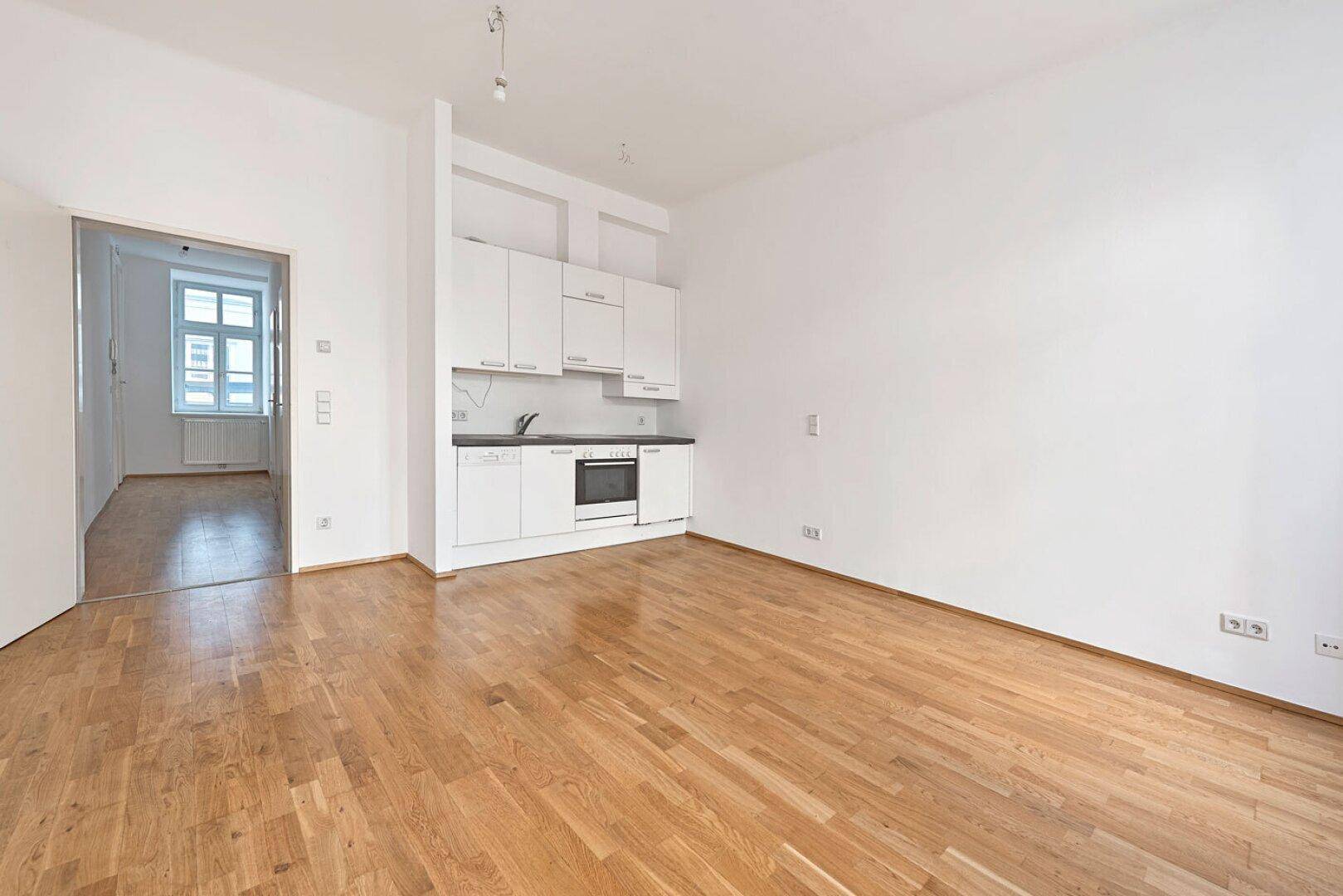 Heller Wohnbereich mit integrierter Küchenzeile, Holzboden und Zugang zu einem weiteren Zimmer.