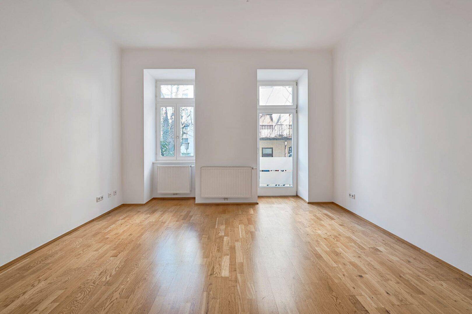 Helles Zimmer mit Holzboden, weißen Wänden und zwei Fenstern, bereit zur individuellen Gestaltung.