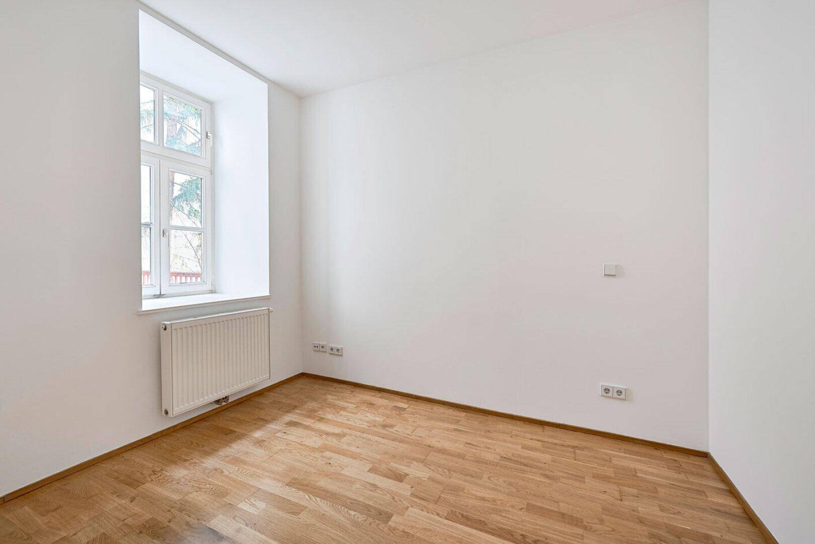 Helles Zimmer mit Holzboden, weißen Wänden und einem Fenster, ideal als Schlaf- oder Arbeitszimmer.