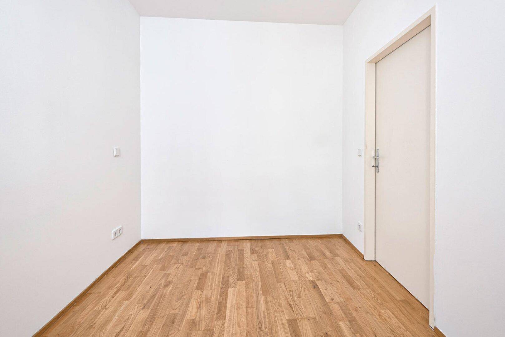 Leerer Raum mit Holzboden und weiß gestrichenen Wänden, eine geschlossene Tür ist sichtbar.