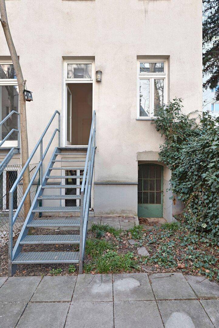 Außenansicht des Gebäudes mit Metalltreppe zum Eingang und begrüntem Bereich.