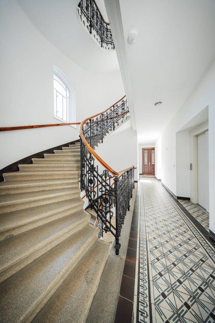 Historisches Treppenhaus mit geschwungenem Geländer und kunstvollem Fliesenboden, inklusive Aufzugzugang.