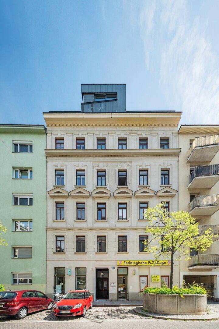 Historische Fassade eines Mehrfamilienhauses mit Geschäften im Erdgeschoss und geparkten Autos.