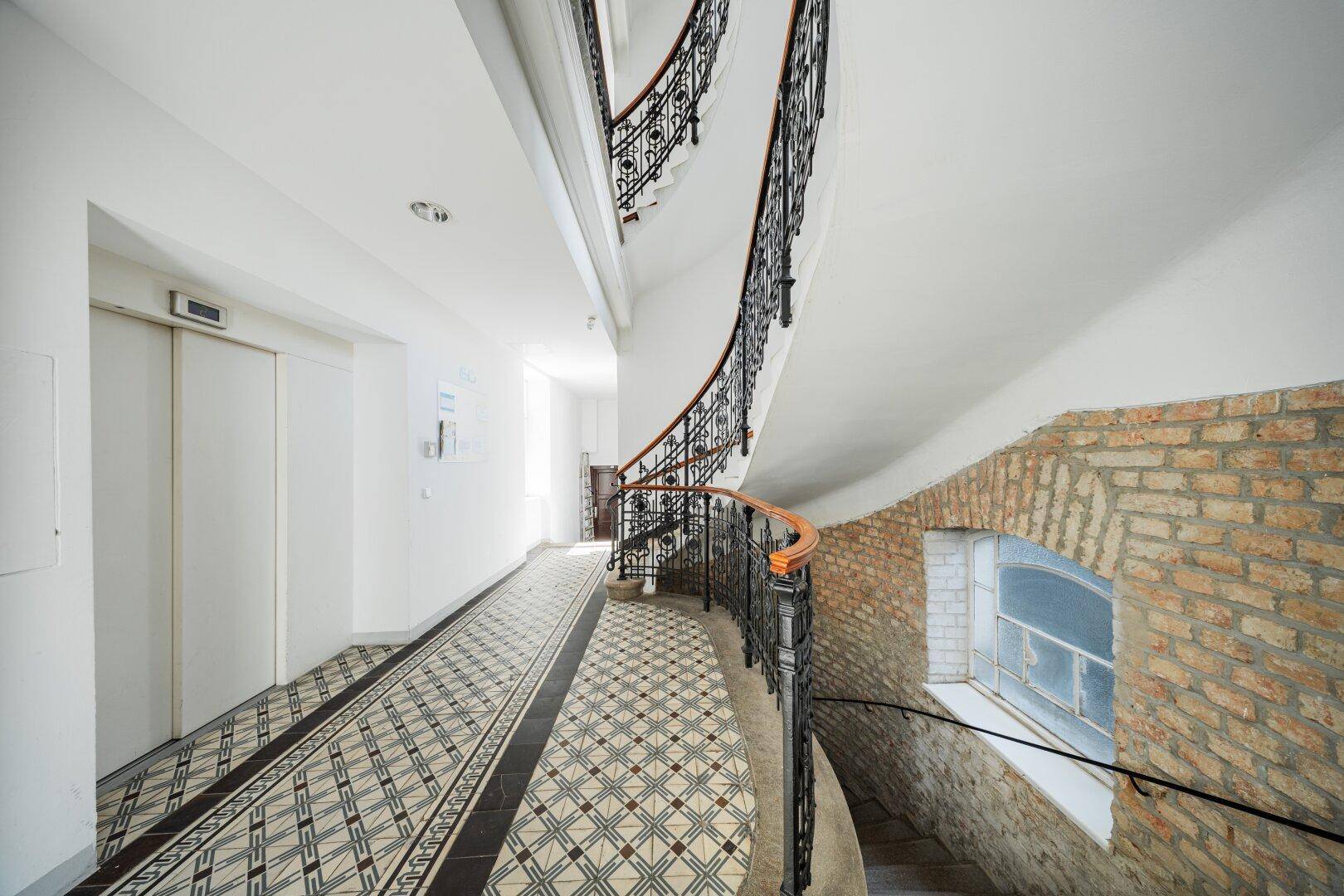 Historisches Treppenhaus mit Aufzug, schmiedeeisernem Geländer und freigelegter Ziegelwand.