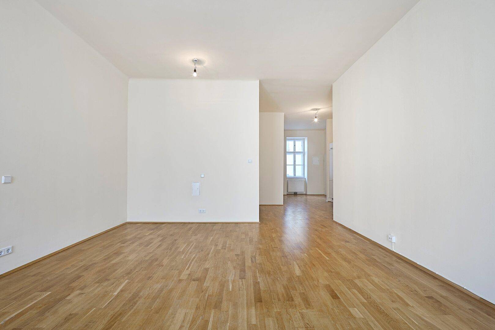Geräumiger Wohnbereich mit Parkettboden und Zugang zu einem hellen Flur mit Fenster.