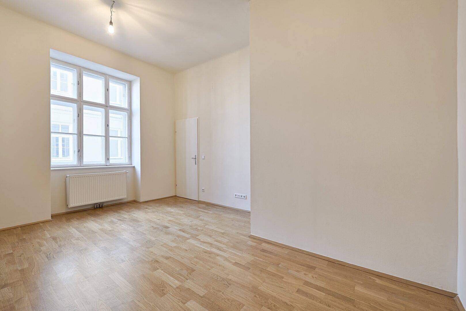 Geräumiges, helles Zimmer mit Parkettboden und einem großen Fenster, ideal für Wohn- oder Schlafbereich.