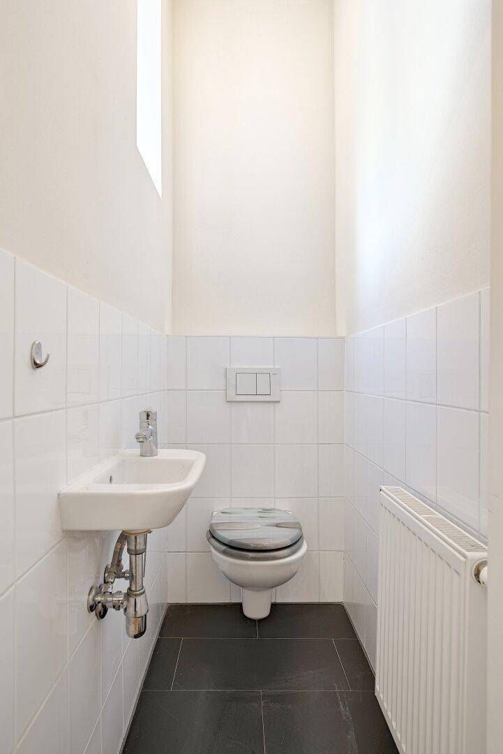 Separates WC mit kleinem Waschbecken, Toilette und Heizkörper, hell gefliest.