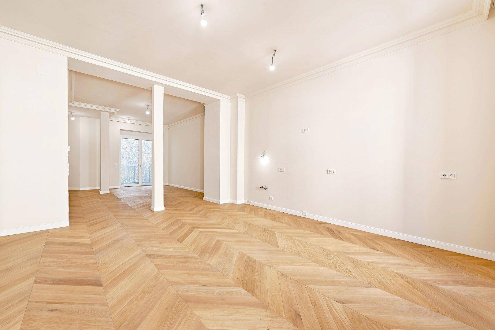 Geräumiges, leeres Wohnzimmer mit elegantem Fischgrätparkett und Blick auf den Balkon.