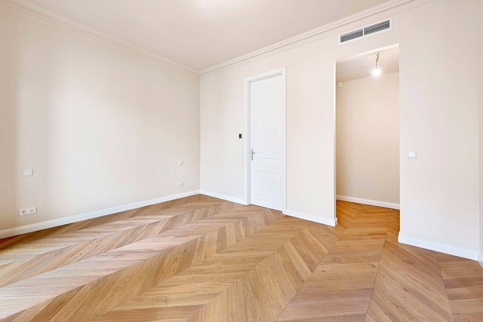 Helles Zimmer mit Fischgrätparkett und Zugang zu einem weiteren Raum, ideal für offene Wohnkonzepte.