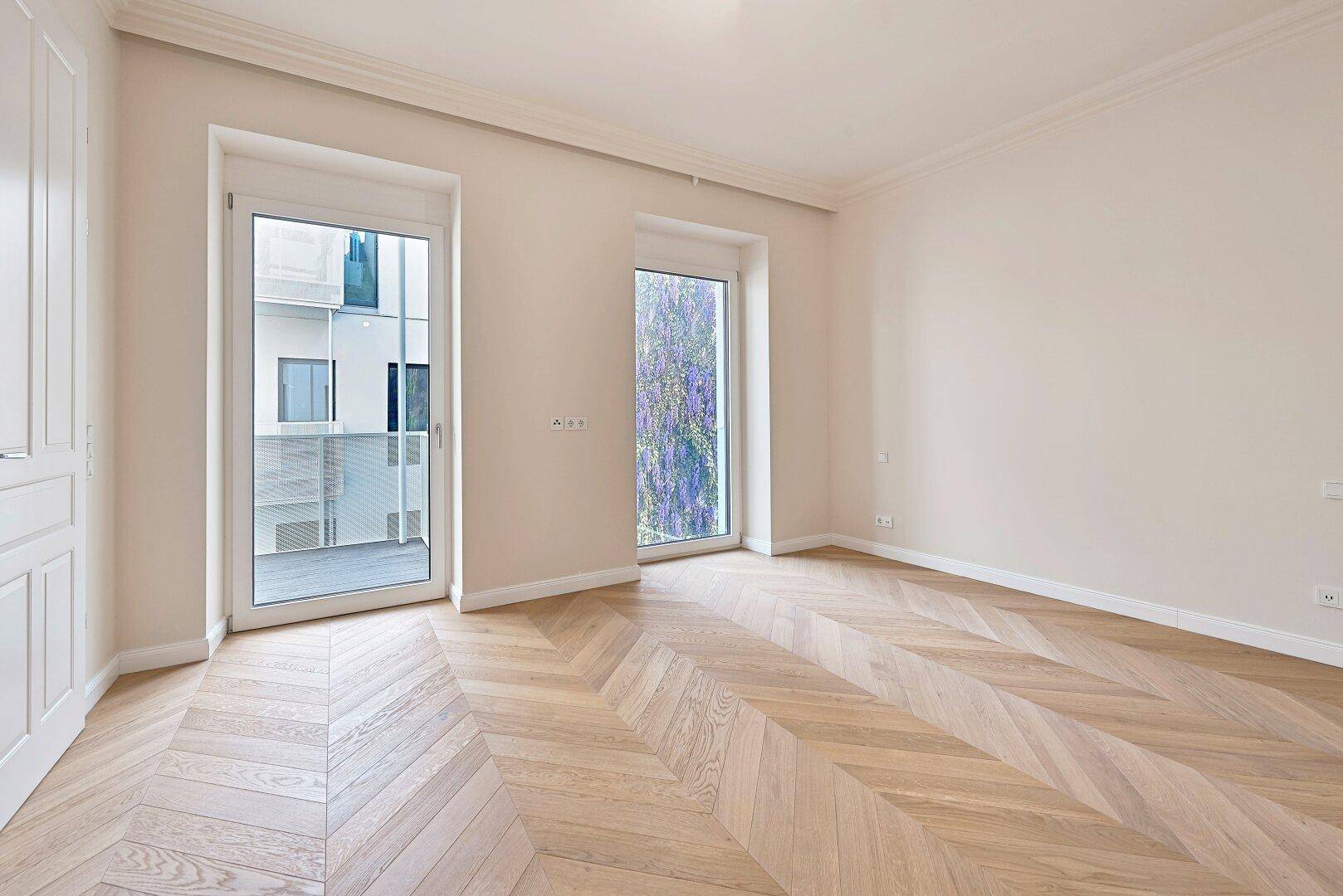 Geräumiges Zimmer mit Fischgrätparkett und großen Fenstern mit Blick auf eine begrünte Wand.