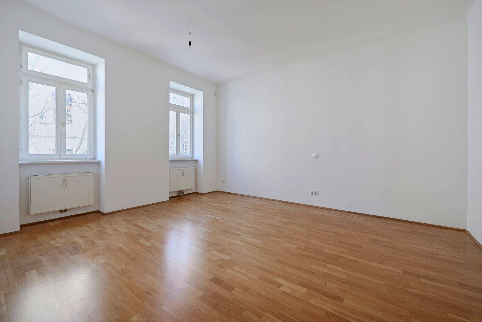 Lichtdurchfluteter Raum mit Parkettboden und zwei großen Fenstern, ideal als Wohn- oder Schlafzimmer.
