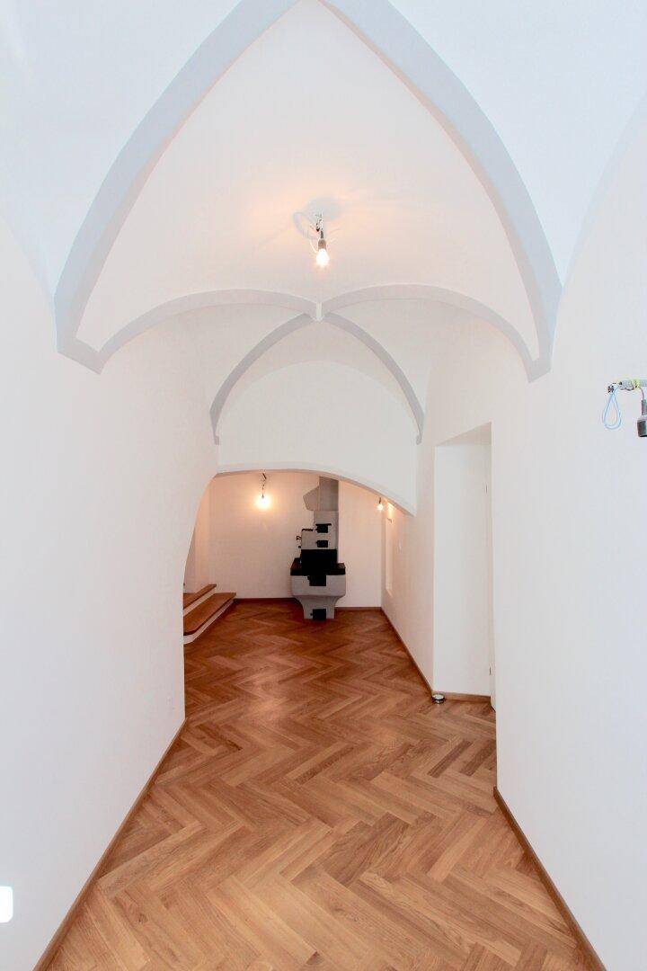Langer Flur mit Fischgrätparkett, Gewölbedecke und einem traditionellen Kachelofen im Hintergrund.