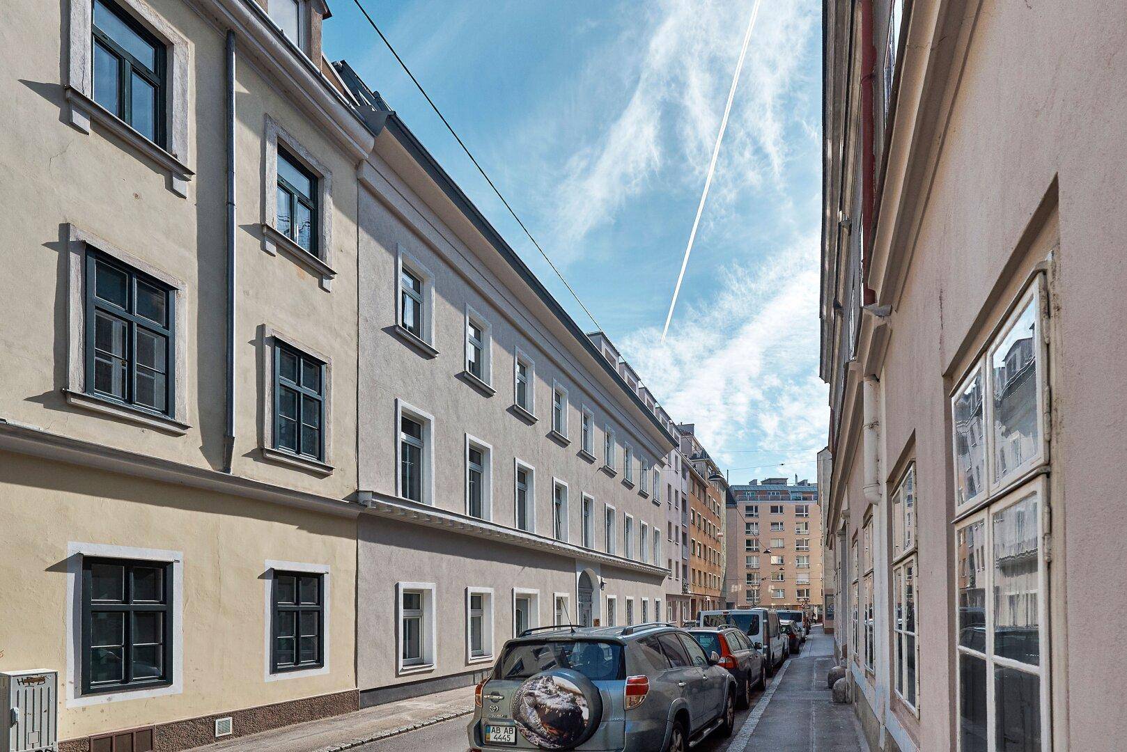 Blick auf eine ruhige Wohnstraße mit geparkten Autos und historischen Gebäudefassaden unter blauem Himmel.