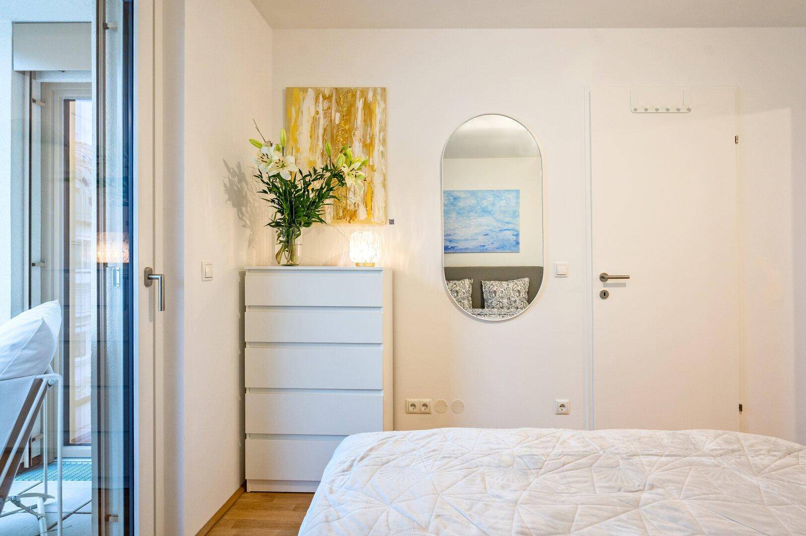 Helles Schlafzimmer mit weißer Kommode, Spiegel und Zugang zum Balkon.