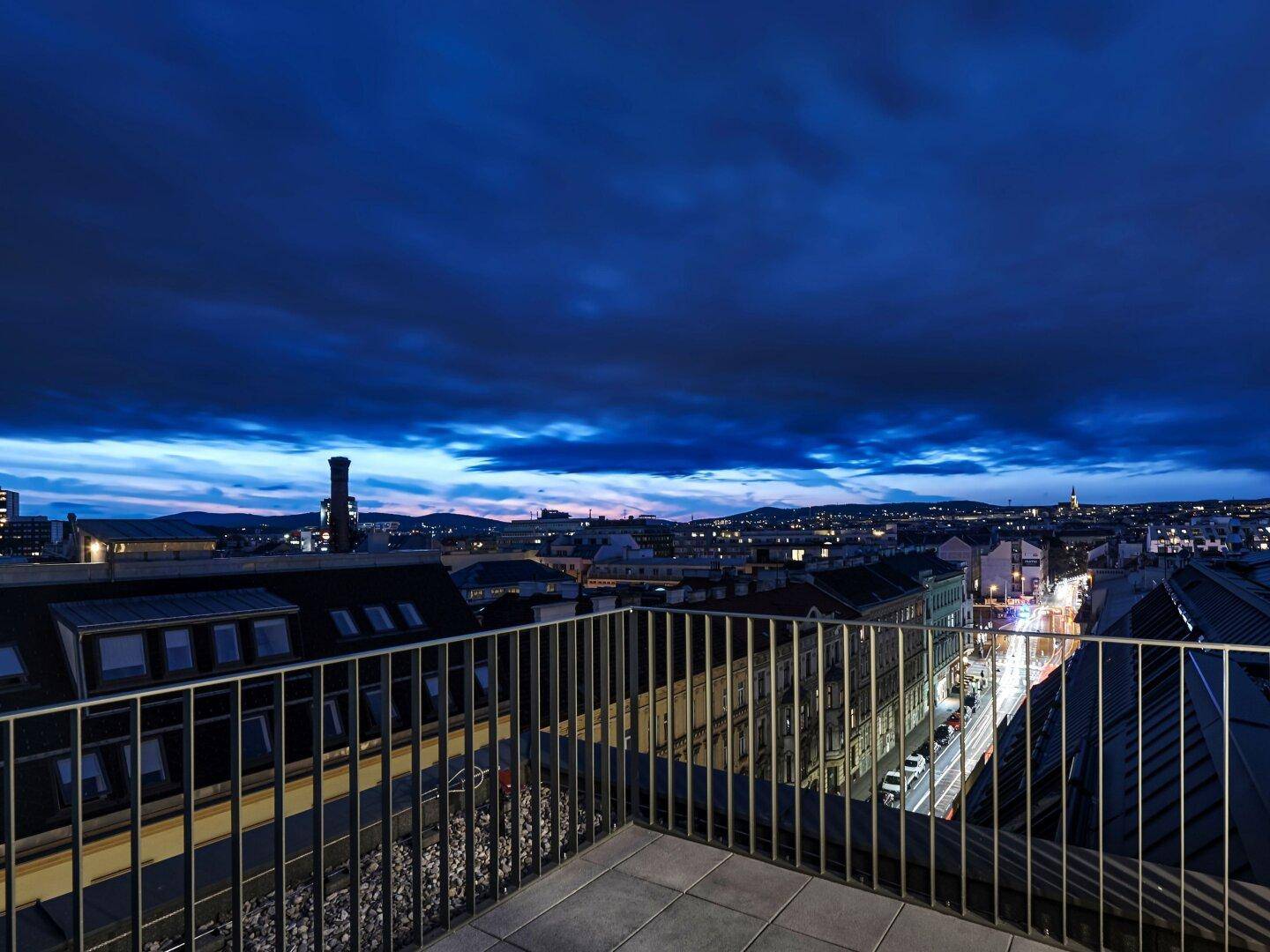 Dachterrasse mit weitem Blick über die Stadt bei Einbruch der Dunkelheit und dramatischem Himmel.