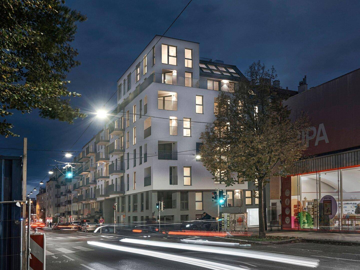 Beleuchtetes modernes Wohngebäude an einer städtischen Kreuzung bei Nacht mit vorbeifahrenden Autos.