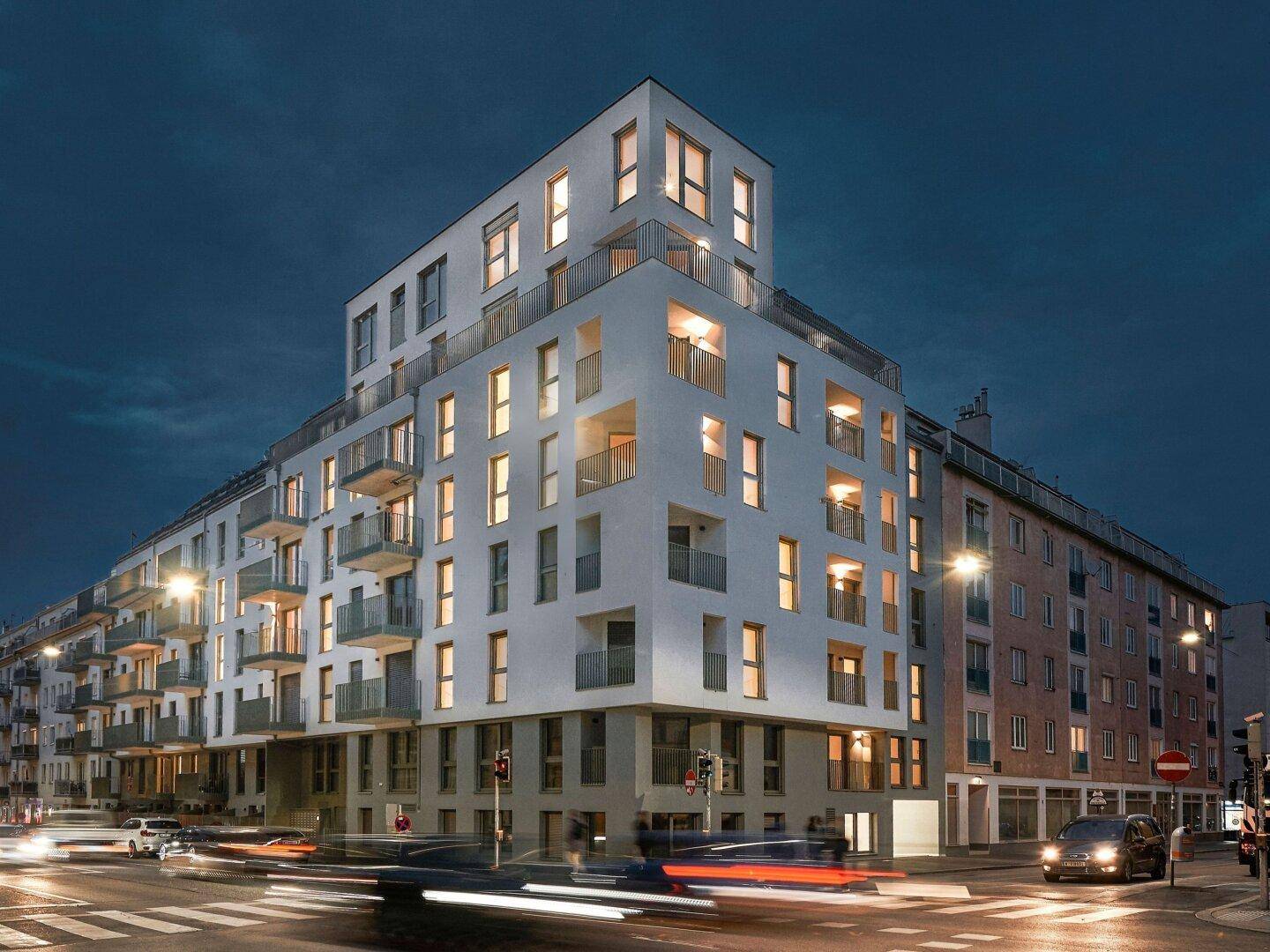 Modernes Wohngebäude mit beleuchteten Fenstern an einer städtischen Kreuzung in der Abenddämmerung.