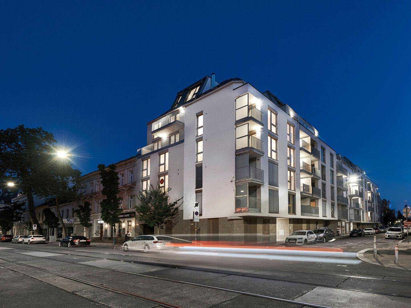 Modernes Eckgebäude mit weißer Fassade, großen Fenstern, Balkonen und Dachterrasse in urbaner Umgebung.